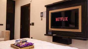 Quarto de hotel com cama, TV na parede exibindo 'NETFLIX', bandeja de madeira com lanches e uma cadeira branca.
