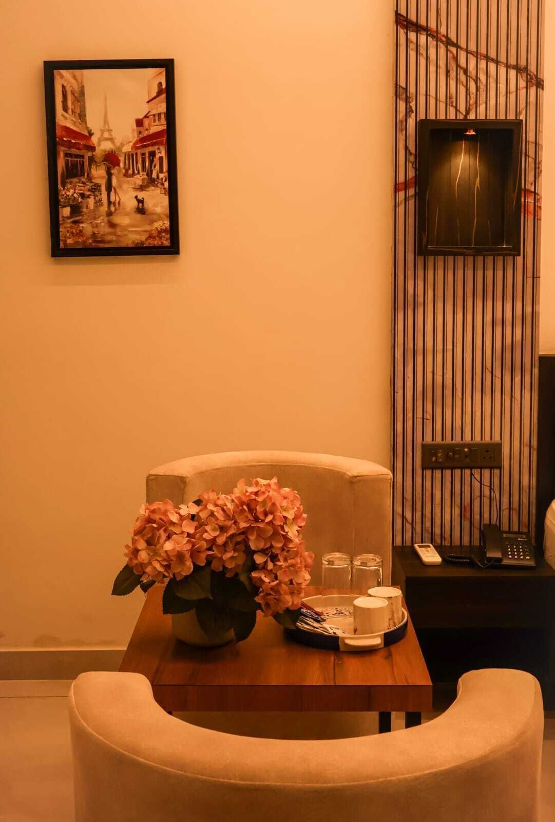 A hotel room with a framed painting, a vase of flowers, a table with a phone and a lamp, and a chair.
