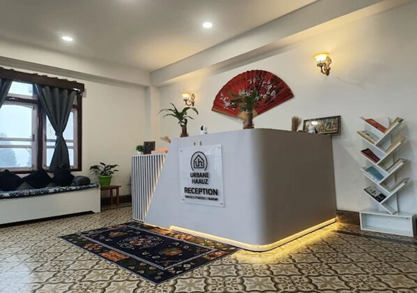 A reception area with a reception desk, a red fan, a potted plant, a bookshelf, and a patterned rug.