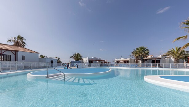 Ein großer, klarer Swimmingpool mit mehreren Bahnen, umgeben von weißen Gebäuden und Palmen.