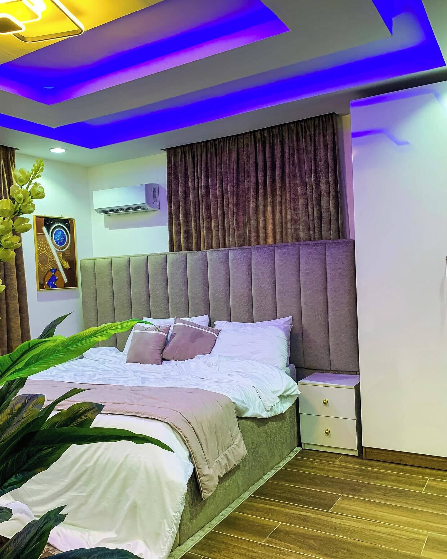 A modern bedroom with a bed, bedside tables, a wall-mounted air conditioner, and a ceiling with blue lighting.