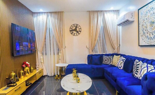 A modern living room with a blue sofa, a round coffee table, a wall clock, and a flat-screen TV mounted on a wooden panel. - ELIZ APARTMENT (Osogbo)