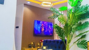 A modern living room with a flat-screen TV, a blue sofa, a decorative plant, and a ceiling light fixture. - ELIZ APARTMENT (Osogbo)