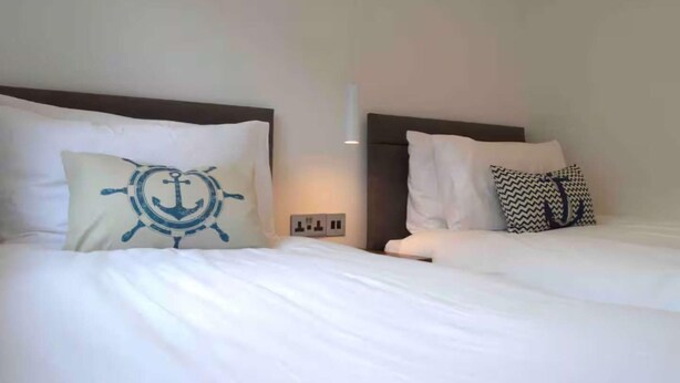 Two beds with white bedding and decorative pillows, a wall-mounted light, and a power outlet. - Riverside House, Lancaster (Lancaster)
