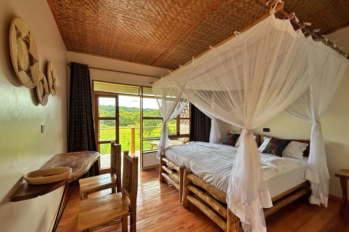 Une chambre avec un lit sous un dais, un bureau en bois, une chaise et une vue sur un paysage verdoyant grâce à de grandes fenêtres.