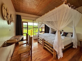 Una habitación con una cama bajo un dosel, un escritorio de madera, una silla y vistas a un paisaje verde a través de amplios ventanales.