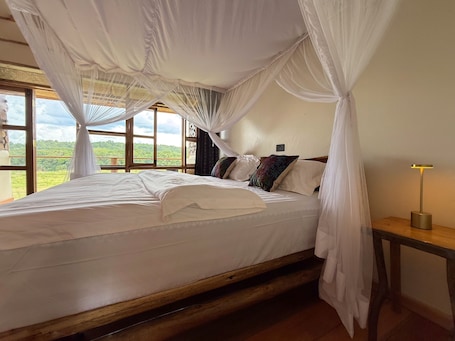 A bedroom with a canopy bed, a bedside table with a lamp, and a view of greenery through large windows.