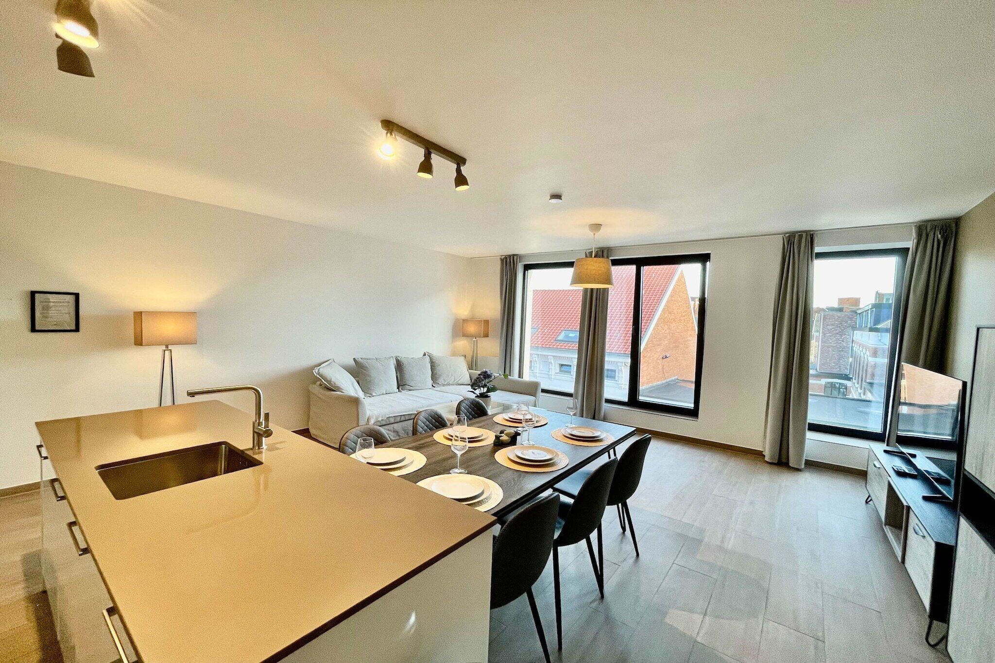 A modern kitchen with a central island, dining area with chairs, a sofa, and large windows with curtains.