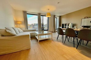 Une chambre d’hôtel moderne équipée d’un canapé, d’une table basse, d’un coin repas avec des chaises, d’une télévision et de grandes fenêtres avec des rideaux.