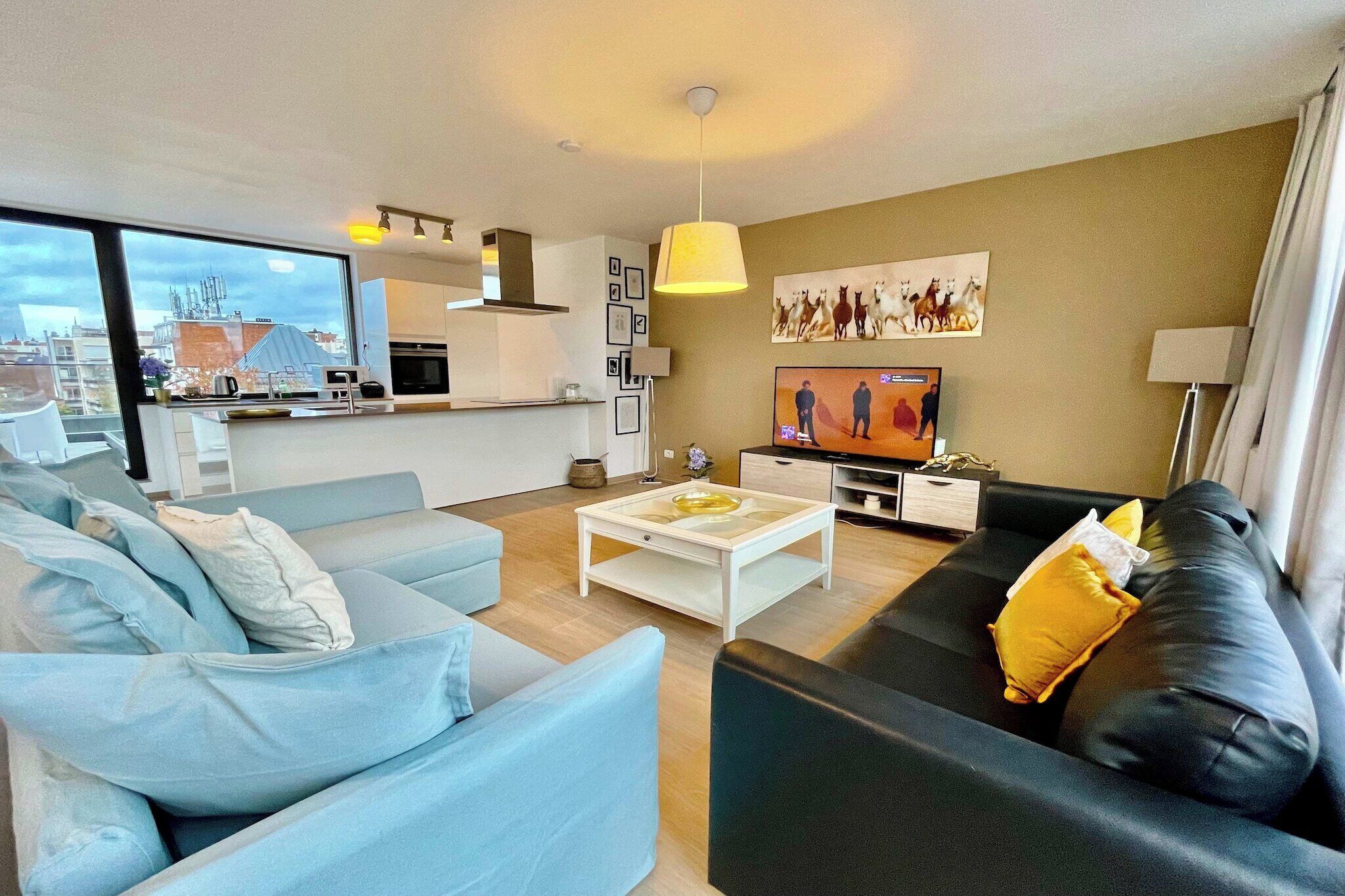 A modern living room with a sofa, a coffee table, a TV, and a kitchen area visible through a large window.