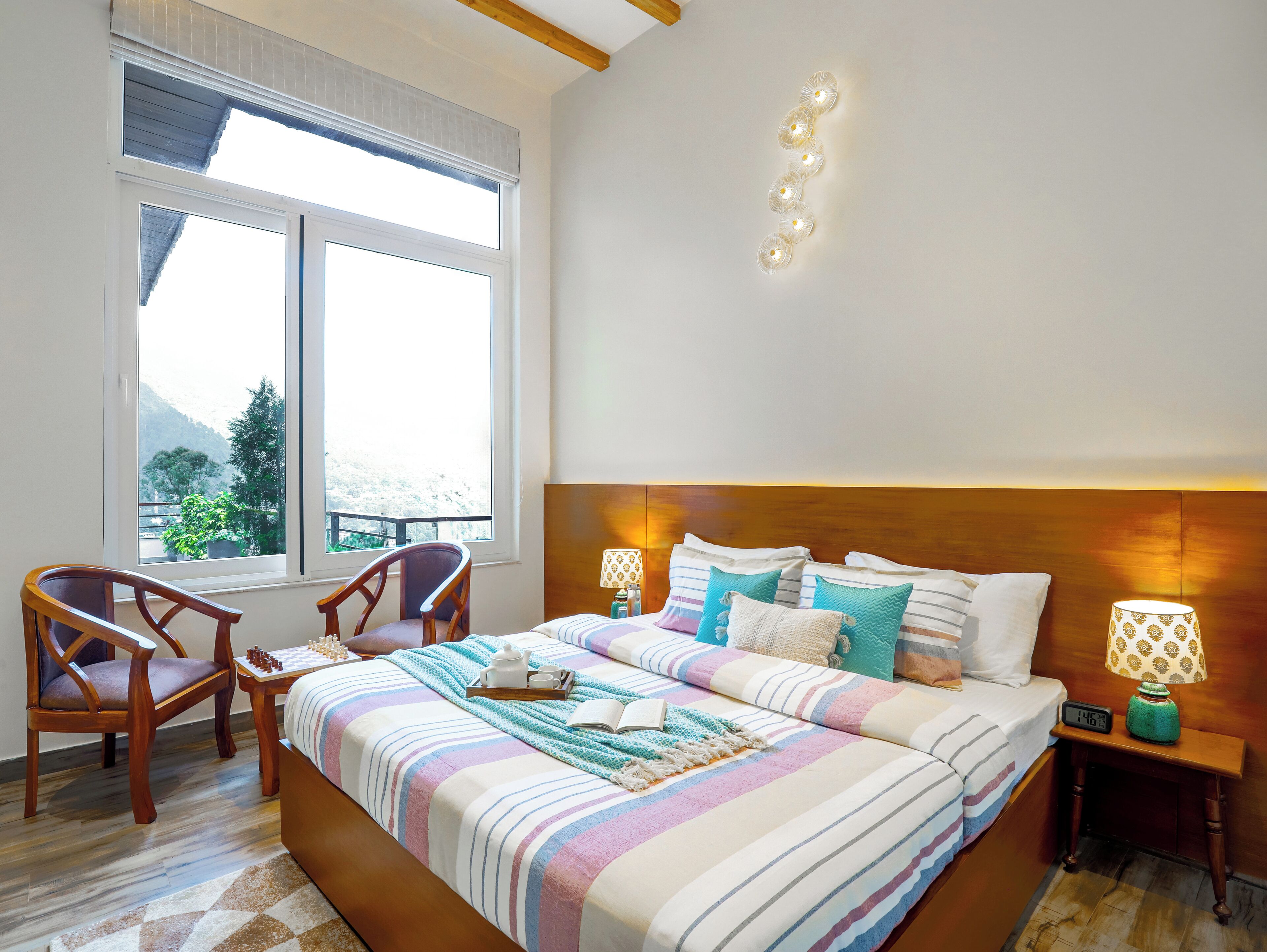 A bedroom with a large bed, a wooden chair, a nightstand with a lamp, and a window with a view of greenery.