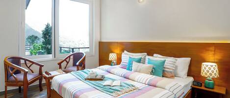 A bedroom with a large bed, a wooden chair, a nightstand with a lamp, and a window with a view of greenery.
