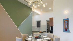 A dining area with a round table set for four, chairs, a chandelier, a wall-mounted artwork, and a kitchen area in the background. - ELIVAAS Antarmann Casa Mirage 3BHK Villa (Bhimtal)