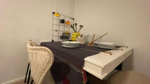 Uma área de refeições com uma mesa posta para dois, uma cadeira, uma estante na parede com objetos e um vaso com flores.