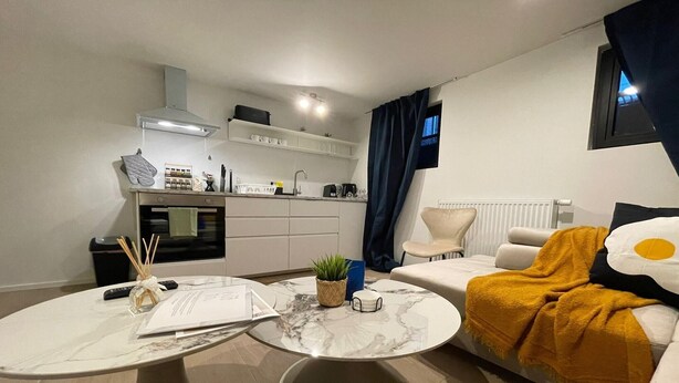 Uma sala de estar moderna com uma mesa de centro redonda de mármore, um sofá com manta amarela mostarda, uma pequena planta em vaso e uma área de cozinha com armários brancos e bancada preta.