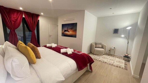 Een slaapkamer met een bed, rode gordijnen, een schilderij, een stoel, een vloerlamp en een nachtkastje.