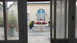 En døråbning der fører til en gårdhave med en springvand, plantekrukker og en bygning med buede vinduer.