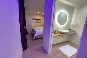 Une salle de bain moderne avec un miroir rond, un lavabo blanc et un lit en arrière-plan.