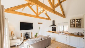 A spacious living area with a high ceiling, wooden beams, a flat-screen TV, a fireplace, a sofa, and a kitchen with white cabinets and wooden countertops. - The Annexe of Richmond Barn - Gateway to Cotswolds (Swindon)