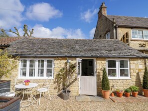 A stone cottage with a tiled roof, a small garden with potted plants, and a white door. - Hallidays Cottages - The Cottage (Oakham)