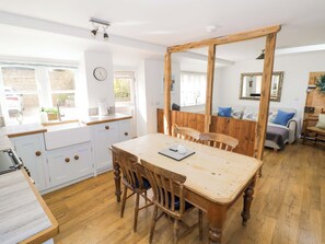 A dining area with a wooden table and chairs, a kitchen with white cabinets, and a large mirror on the wall. - Hallidays Cottages - The Cottage (Oakham)