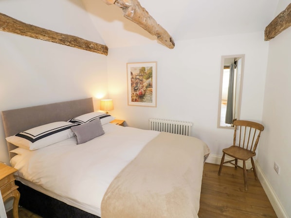 A bedroom with a bed, a nightstand, a lamp, a radiator, a window, and a wooden chair. - Hallidays Cottages - The Cottage (Oakham)
