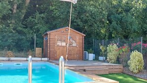 Eine Holzterrasse mit Pool, eine Holzhütte und ein eingezäunter Gartenbereich.