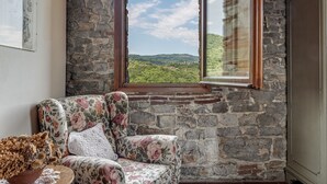 A room with a stone wall, a floral armchair, a view of green hills, and a window with curtains. - Spacious Panoramic Apartment with Pool (Pontassieve)