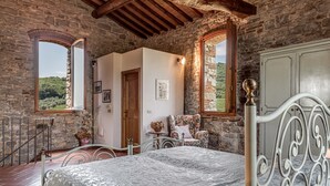 A rustic bedroom with stone walls, a bed, a chair, and a view of a mountainous landscape. - Spacious Panoramic Apartment with Pool (Pontassieve)