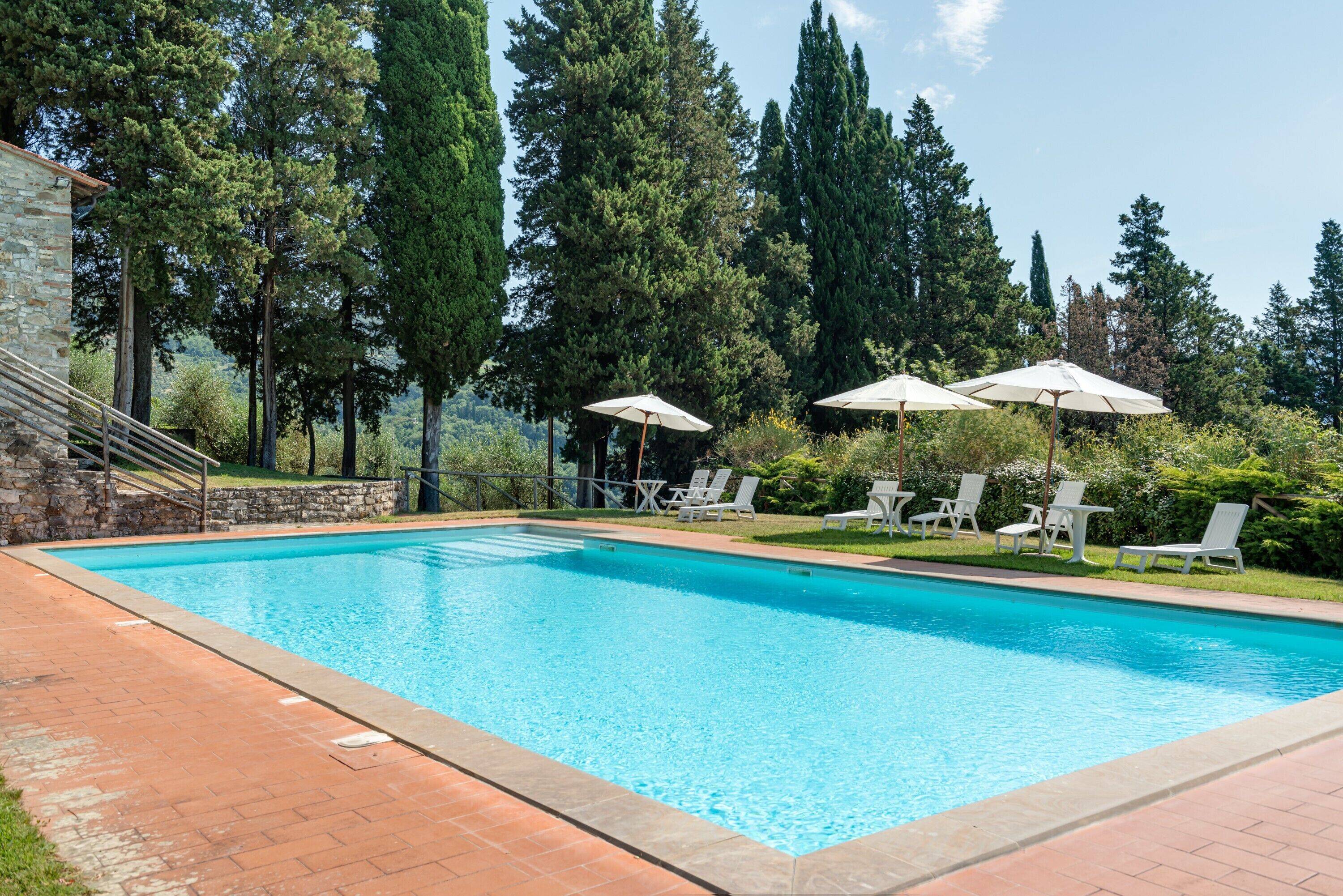 A swimming pool with a stone wall, surrounded by trees and white lounge chairs.