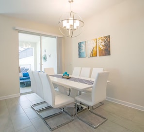 A dining area with a white table, chairs, and a chandelier. There are artworks on the wall and a view of a balcony with blue chairs. - New Listing-Modern Storey Lake Pool Villa- Disney. (Kissimmee)