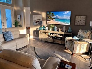 Een woonkamer met een grote flatscreen televisie, een bank, een fauteuil, een salontafel en een houten dressoir met diverse spullen.