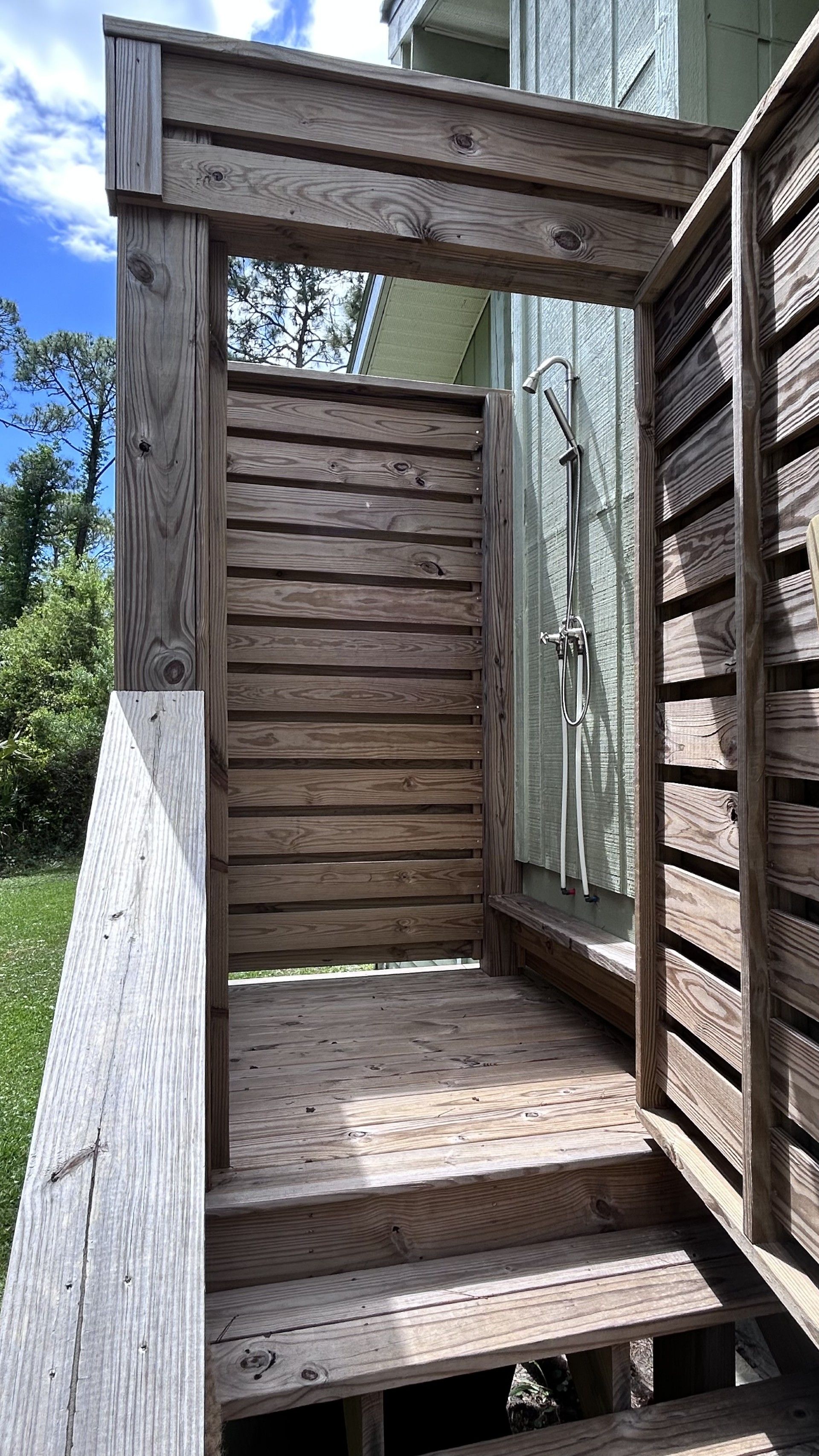 A wooden outdoor shower structure with steps leading up to it, a metal showerhead, and a wooden wall.