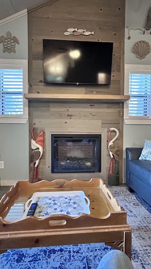 A living room with a fireplace, a flat-screen TV mounted above it, and a blue couch. - Breezy Bay Cabin – A Cozy Bayfront Retreat for Couples & Small Families (Cape San Blas)