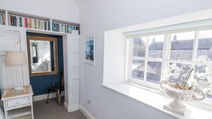 A room with a bookshelf, a mirror, a window with a view of houses, and a white vase with flowers. - Hallidays Cottages - The Folly (Oakham)