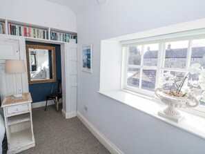 A room with a bookshelf, a mirror, a window with a view of houses, and a white vase with flowers. - Hallidays Cottages - The Folly (Oakham)