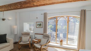 A room with wooden flooring, a sofa, two armchairs, a small table, a radiator, a window with curtains, and a view of the outside. - Hallidays Cottages - The Folly (Oakham)