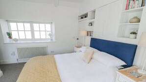 A neatly arranged bedroom with a bed, bedside tables, a radiator, a window, and a bookshelf. - Hallidays Cottages - The Folly (Oakham)