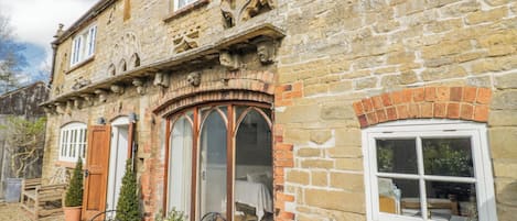 Un edificio de piedra con un amplio ventanal arqueado, una ventana más pequeña y una puerta. Hay una mesa con sillas y plantas en macetas en el exterior.