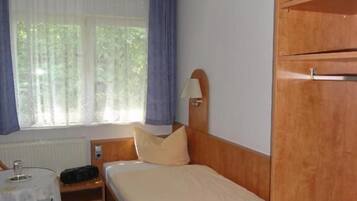 A bedroom with a wooden bed, a bedside table with a lamp, a small desk with a chair, and a window with curtains.