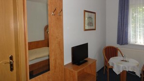 A room with a wooden wardrobe, a television on a small table, a chair, and a window with curtains.