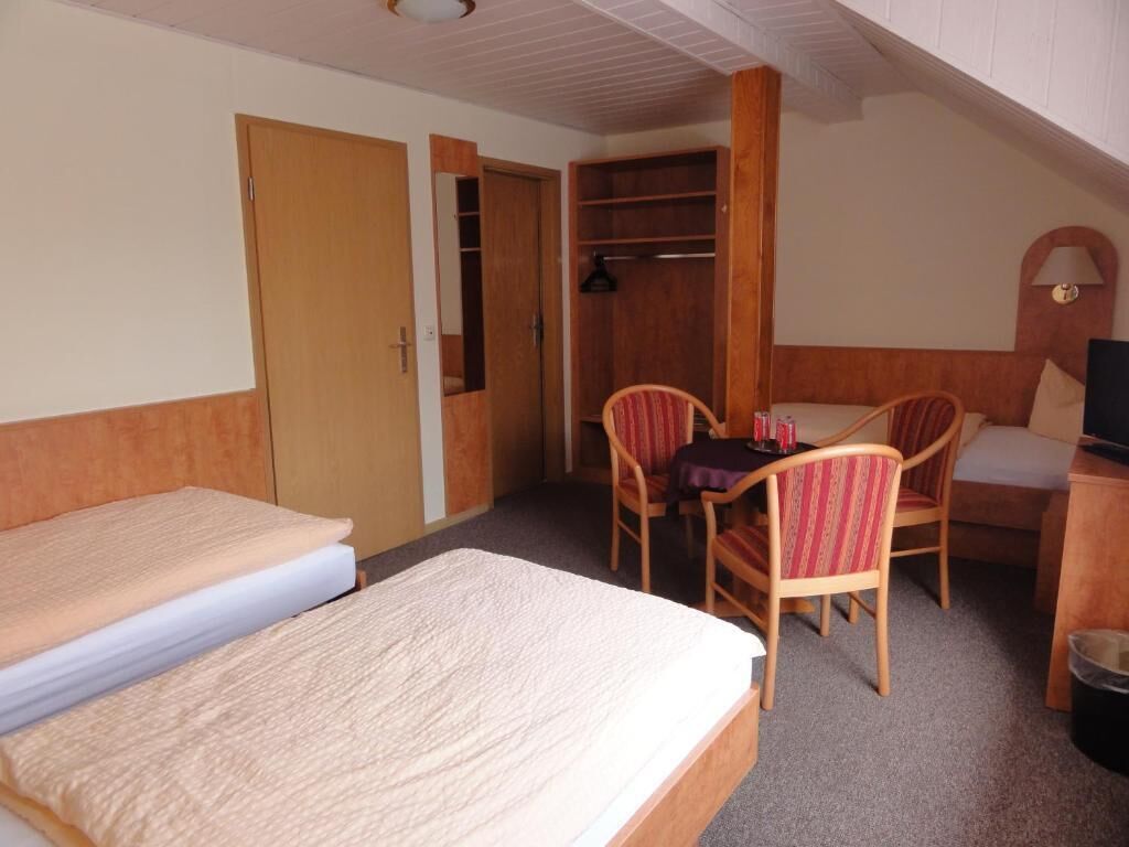 A hotel room with two beds, a wooden wardrobe, a desk with a chair, and a small table.