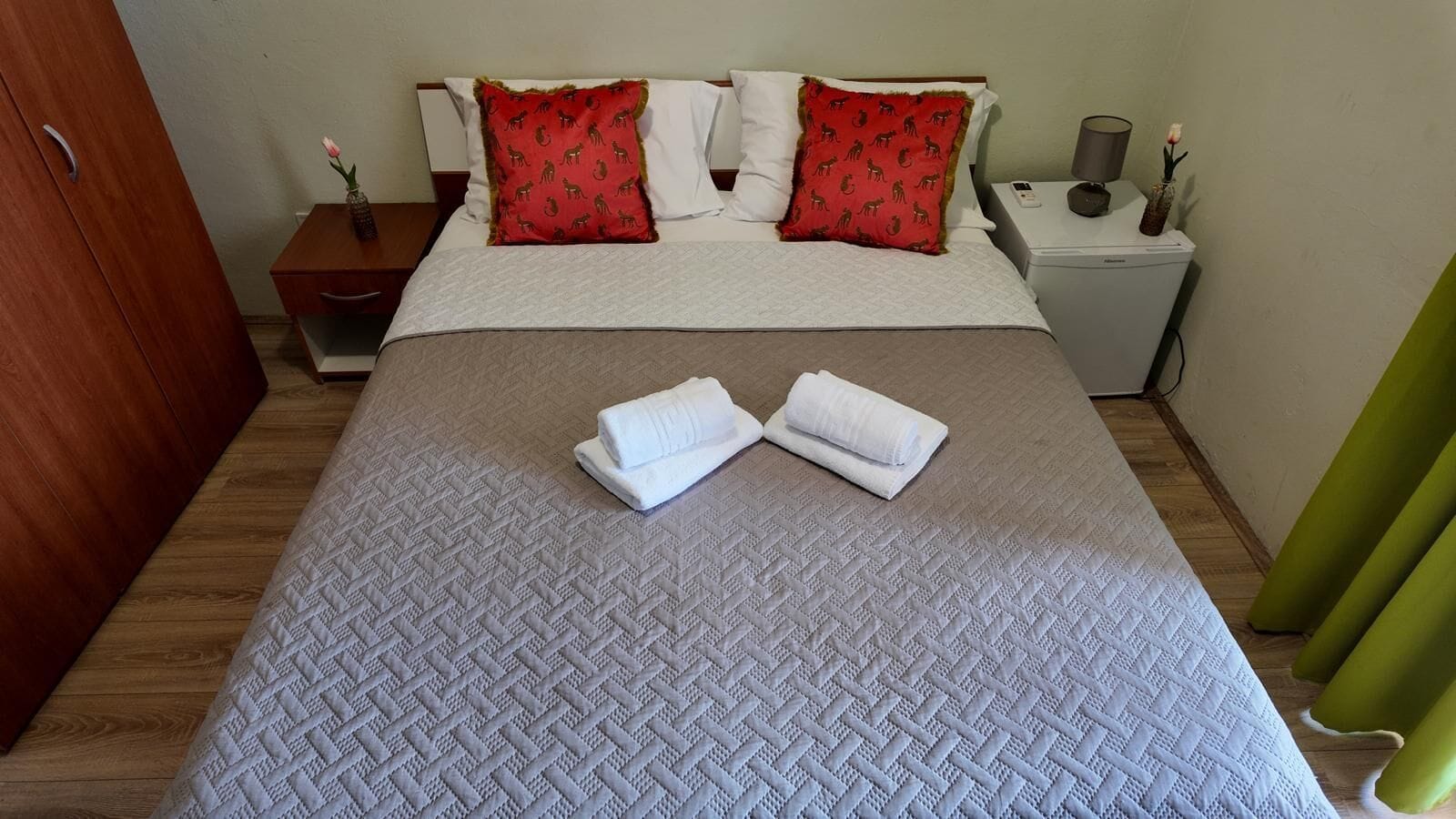 A neatly made bed with white linens, two red pillows with animal prints, a wooden nightstand with a lamp, and a green curtain.
