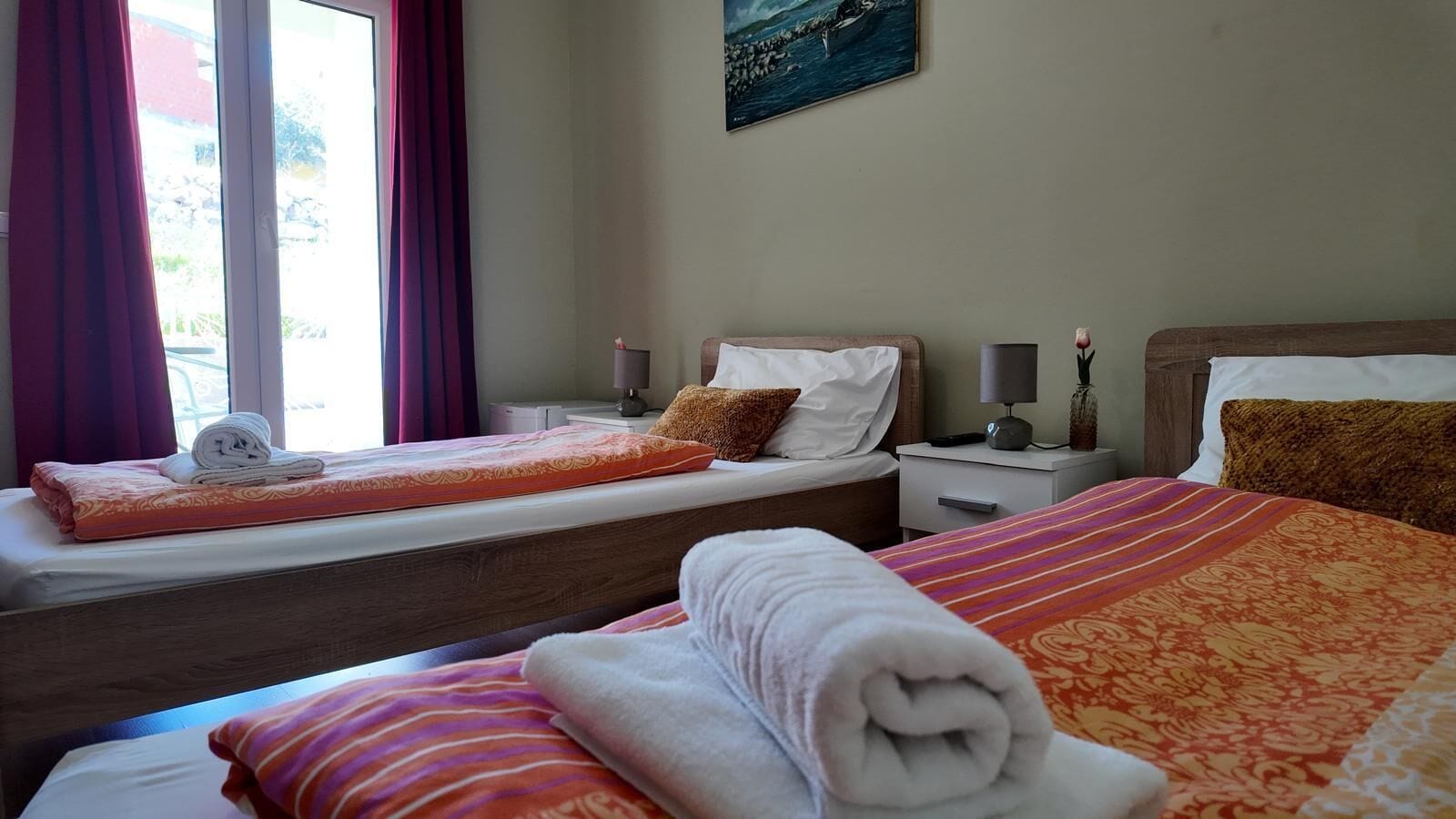 A hotel room with two beds, a window with curtains, a bedside table with a lamp, and a painting on the wall.