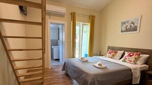 A bedroom with a bed, a wooden ladder, a bathroom, and a window with curtains. - Adrian's Beach House by Max Gastro (Marina)