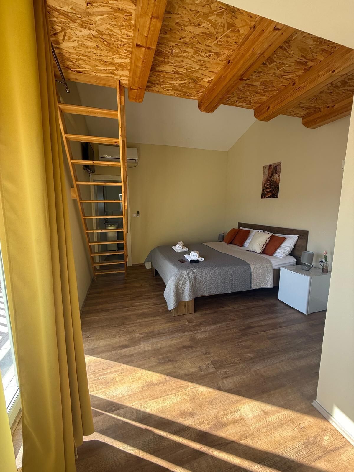 A bedroom with a wooden ceiling, a bed, a nightstand, a desk, a ladder, and a window with curtains.