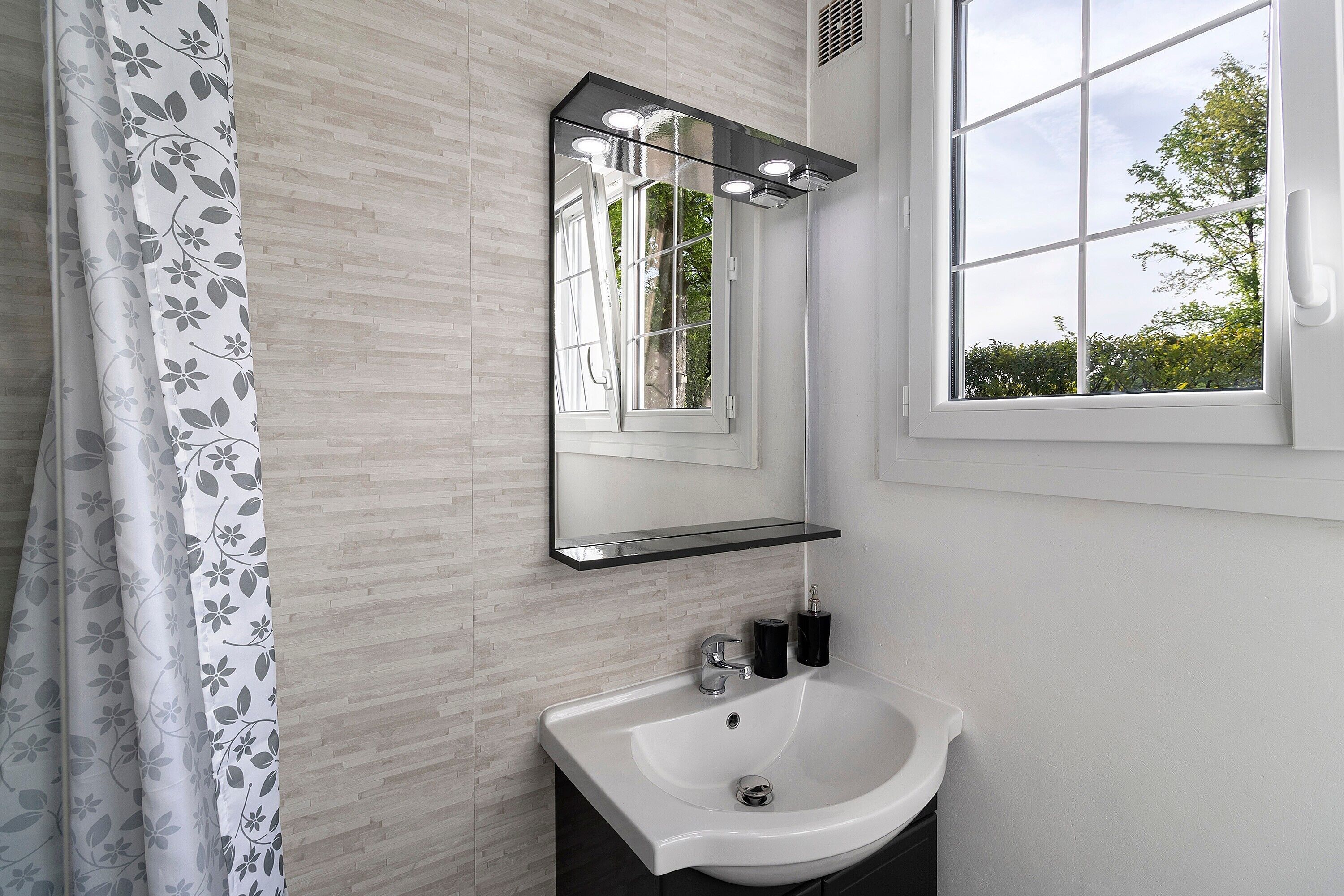 A bathroom with a white sink, a mirror with lights, and a window with a view of trees.