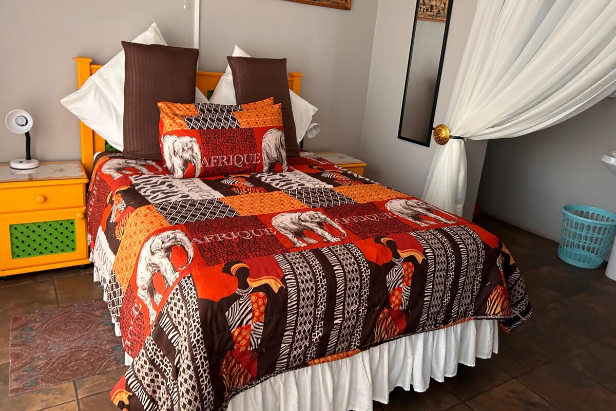 A bed with a patterned bedspread, pillows, a bedside table with a lamp, and a wall-mounted mirror.