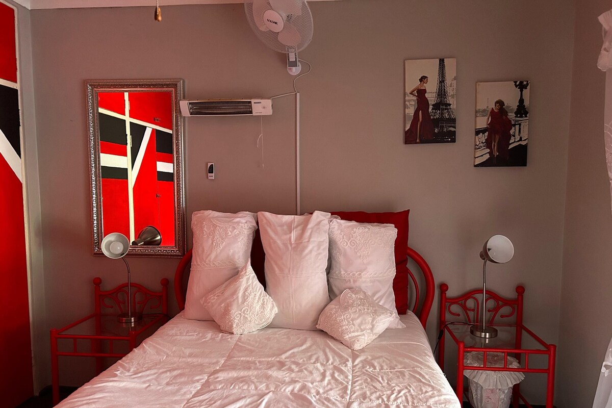 A bedroom with a red bed frame, white bedding, a red-framed mirror, a wall-mounted air conditioner, and two wall-mounted lamps.