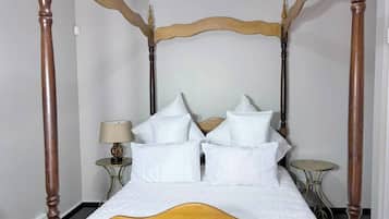 A four-poster bed with white bedding and multiple pillows, a bedside table with a lamp, and a small round table.